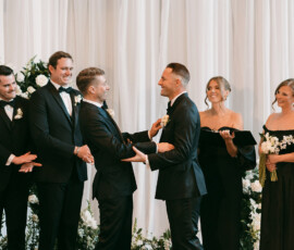 Two grooms hold hands and smile at their Louisiana wedding, surrounded by their formally dressed wedding party and floral decor.
