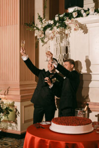 Two men in suits toast with champagne and smile beside a raspberry-topped cake at a New Orleans wedding reception.