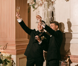 Two men in suits toast with champagne and smile beside a raspberry-topped cake at a New Orleans wedding reception.