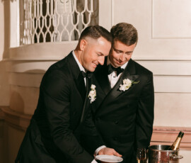 Two men in tuxedos cut a raspberry cake at a festive table with champagne, flowers, and Louisiana wedding inspiration.