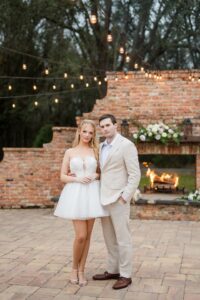Couple poses by a flower-adorned brick fireplace with string lights at a romantic Gulf South wedding.