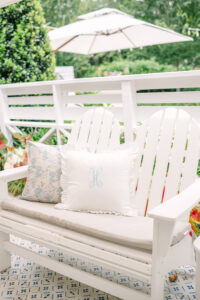 White wooden bench with beige cushion and pillows, one with H monogram, on a patio with greenery and large umbrella in background.