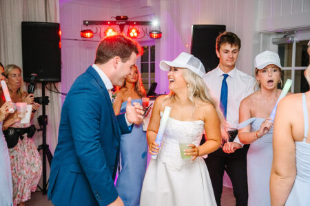 Bride in white dress and hat laughs with man in blue suit at a lively New Orleans wedding reception with glow sticks and drinks.