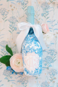Blue and white floral wine bottle with a white bow on a floral surface, surrounded by pink roses and green leaves—wedding décor.