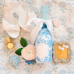 Elegant wedding accessories—white bow heels, floral blue bottle, flowers, gold perfume, ring, and pearls on a floral background.