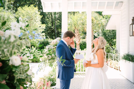 Bride laughs as groom wipes his eyes holding a note on porch, surrounded by vibrant greenery at New Orleans wedding.