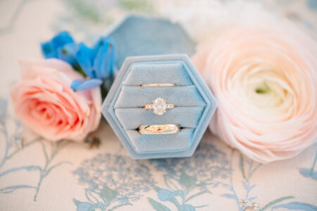 Gold rings in a blue hexagonal box, surrounded by pink rose, ranunculus, and blue blooms on a floral-patterned surface.
