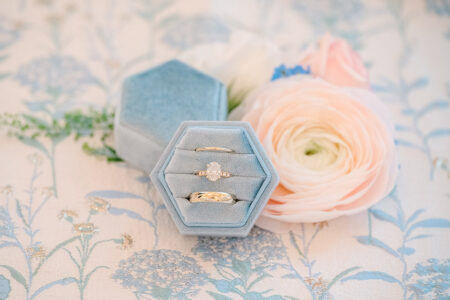 Blue velvet hexagonal ring box with diamond and gold rings beside a pink ranunculus on floral fabric at a Louisiana wedding.