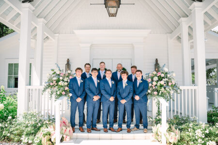 Groomsmen in blue suits smile on steps of a white flower-adorned building, classic Louisiana wedding inspiration.