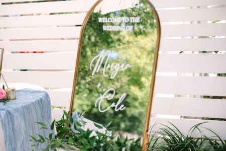 Gold-framed mirror sign says “Welcome to the wedding of Megan & Cole” with greenery, white fence, and blue-draped table behind.