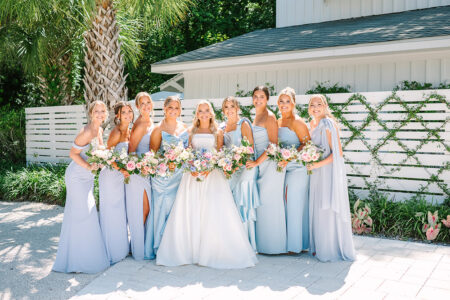 Nine women in blue and white dresses hold flower bouquets and smile outside, with greenery and a white building behind them.