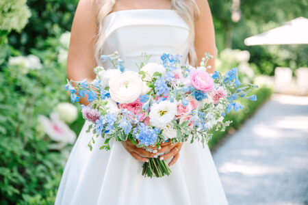 Bride in white dress holding pastel bouquet amid greenery on sunny path, evoking a classic New Orleans wedding.