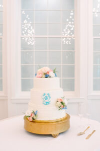 Three-tiered white wedding cake with pink and blue flowers on gold stand, flanked by champagne flutes and glowing window lights.