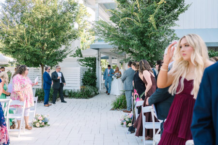 Bride walks outdoors down the aisle with a man, guests watching, greenery and white buildings in classic Louisiana wedding style.