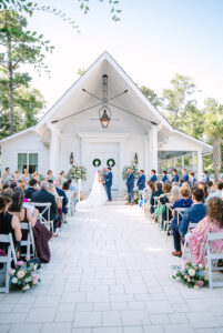 A couple weds at a white chapel altar, surrounded by guests, flowers, and greenery—ideal for classic Gulf South weddings.