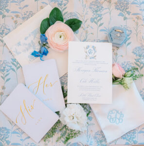 Wedding stationery flat lay with vow books, invitation, ring box, flowers, and handkerchief on a floral background.