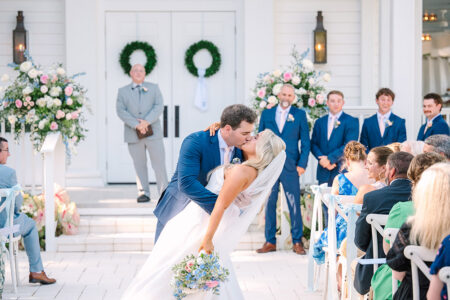 Groom in blue suit dips and kisses bride in white dress at outdoor Gulf South wedding, with guests and smiling groomsmen behind.