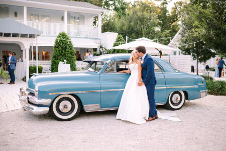Bride and groom kiss by a vintage blue car outside a white house, classic Gulf South wedding scene with guests and greenery.