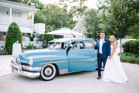 Bride and groom smile by a vintage blue car outside a white house, greenery all around, on a sunny Gulf South wedding day.