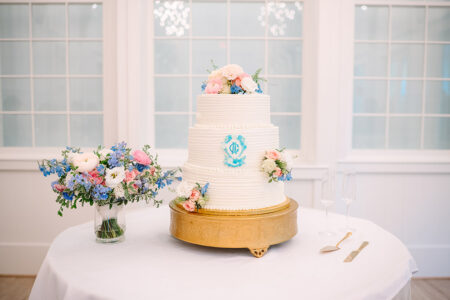 Elegant white three-tier cake with pink and blue flowers on gold stand beside champagne flutes, utensils, and flowers.