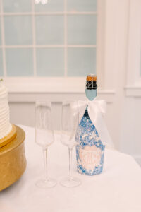 Champagne bottle with white bow, monogrammed flutes, and white cake on gold stand—elegant Louisiana wedding inspiration.