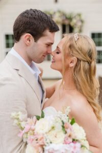 Bride and groom embrace outdoors, smiling with foreheads touching, in front of a white building with pastel bouquet.