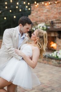 Couple dances by a brick fireplace under string lights at a Louisiana wedding; man in beige suit, woman in strapless white dress.