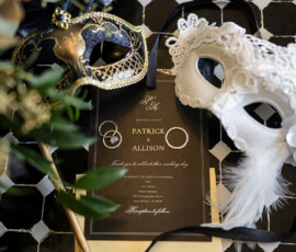 Black and gold wedding invitation suite styled with masquerade masks, wedding bands, and elegant ribbon details on a patterned tile floor.