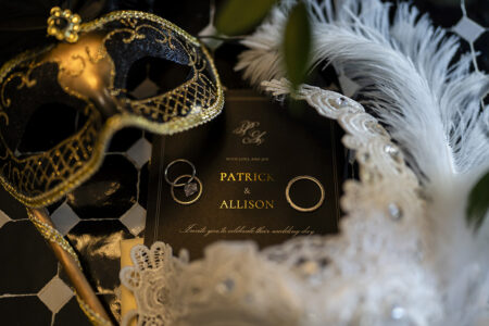 Gold and black masquerade mask, rings, feather, and lace frame a black Patrick & Allison wedding invitation.