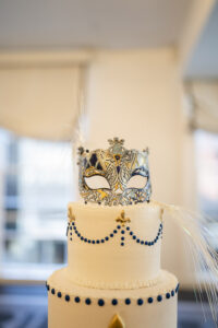 White tiered cake with blue and gold accents, topped with a masquerade mask and feathers for a New Orleans wedding.