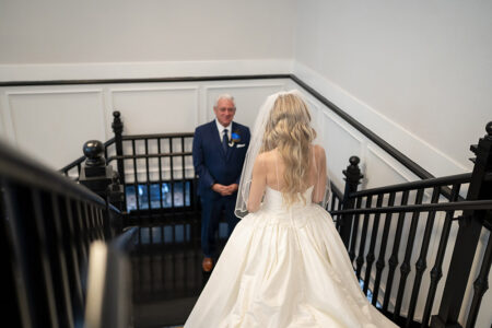 Bride in white gown stands atop staircase, facing older man in blue suit. White paneled walls and black railings indoors.