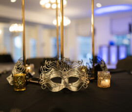 Silver masquerade masks and glowing candle votives decorate black linen reception tables with tall gold centerpieces at an elegant evening wedding reception.