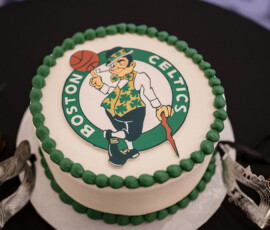 Groom’s cake decorated with the Boston Celtics logo displayed alongside ornate silver masquerade masks and candlelight accents.