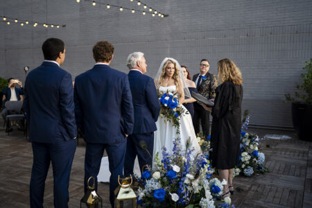 Bride in white gown with blue and white bouquet stands beside older man at outdoor New Orleans wedding, surrounded by blue-suited attendants.