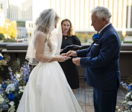 Bride and groom exchange rings during an intimate rooftop wedding ceremony surrounded by lush blue and white floral arrangements.