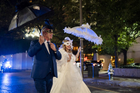 Bride and man in masks with decorated umbrellas walk at night amid city lights, evoking a magical New Orleans wedding.