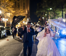 Bride and groom wearing ornate masquerade masks and carrying decorative parasols lead a lively second line parade through downtown New Orleans at night.