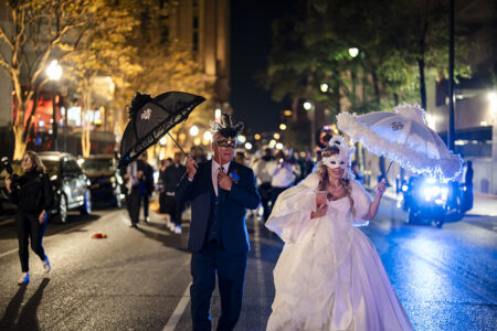 Bride and groom wearing ornate masquerade masks and carrying decorative parasols lead a lively second line parade through downtown New Orleans at night.