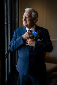 Older man in blue suit smiles by a window, adjusting boutonniere in softly lit room—elegant Gulf South wedding inspiration.