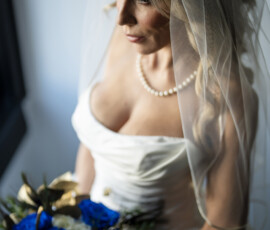 Bride in an off-the-shoulder satin wedding gown and pearl necklace holds a bouquet of vivid blue roses beneath a softly draped veil in an editorial bridal portrait.