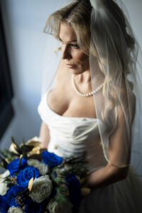 Bride in an off-the-shoulder satin wedding gown and pearl necklace holds a bouquet of vivid blue roses beneath a softly draped veil in an editorial bridal portrait.