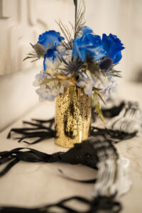 Blue, white, and gold floral arrangement with peacock feathers displayed in a metallic gold vase beside black masquerade masks.
