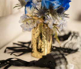 Blue, white, and gold floral arrangement with peacock feathers displayed in a metallic gold vase beside black masquerade masks.