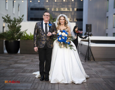 Bride in white gown with blue and white flowers and man in patterned suit smile on New Orleans rooftop wedding.