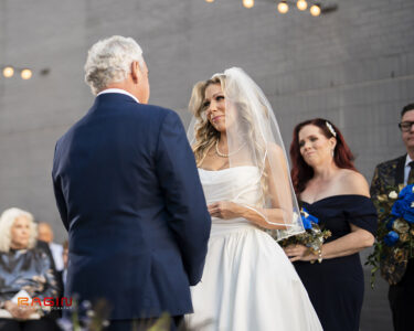 Bride in white dress and veil faces groom in navy suit at outdoor wedding, with bridesmaids and guests watching.