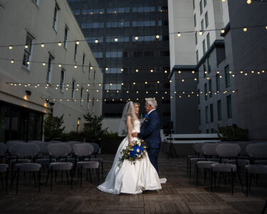 Bride and groom share a quiet moment alone on a rooftop wedding terrace framed by café lights and modern city buildings.