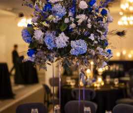 Tall reception centerpiece overflowing with blue roses, hydrangeas, white blooms, and peacock feathers rises above candlelit reception tables.