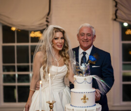 Bride and groom smile beside their elegant white wedding cake decorated with blue beading, gold fleur-de-lis accents, and masquerade-inspired details.