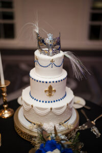 Three-tier white wedding cake adorned with blue bead detailing, gold fleur-de-lis accents, draped icing details, and a dramatic masquerade mask topper with feathers.