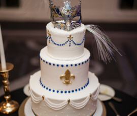 Three-tier white wedding cake adorned with blue bead detailing, gold fleur-de-lis accents, draped icing details, and a dramatic masquerade mask topper with feathers.